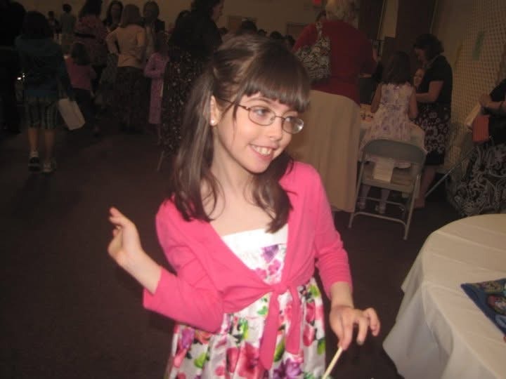 A young girl with glasses who is wearing a pink sweater and a white floral dress at a Girl Scout tea party event.