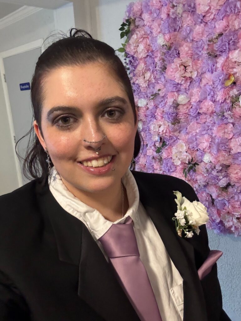 An image of the author wearing a black tuxedo, white button-up, a wisteria tie, and a boutonniere at their best friend’s wedding. Their facial expression is neutral, even though they were ecstatic.