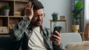A man sitting on a sofa checks his smartphone while holding his head, appearing concerned or frustrated | Photo: Shutterstock