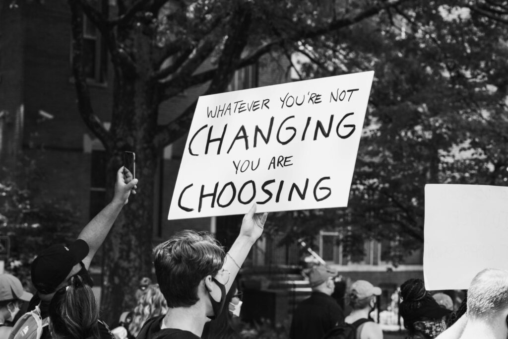 Black and white image of a person holding a sign in a protest. The sign says, “Whatever you’re not changing, you are choosing.”