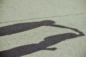 Two shadows, cast long on the pavement are connected as the first pulls the second forcefully by her arm. They are arguing.