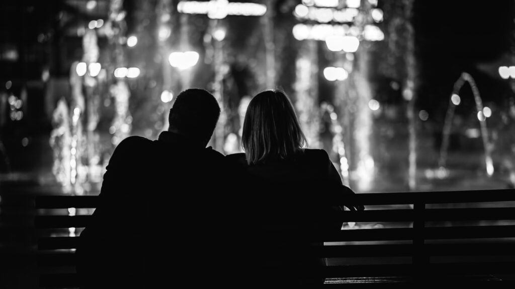 Image of a couple sitting on a bench, looking at a fountain, in black and white.