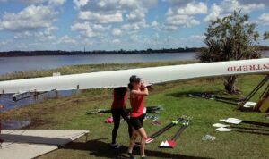 A fifteen/sixteen year old Sophie carries a four-person boat in from the water with a teammate behind her.