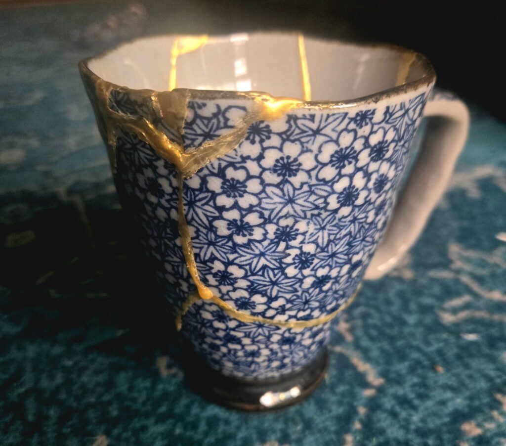 Image of the repaired mug. The mug is light blue with a dark blue floral pattern. Gold resin fills the cracks that were once there.