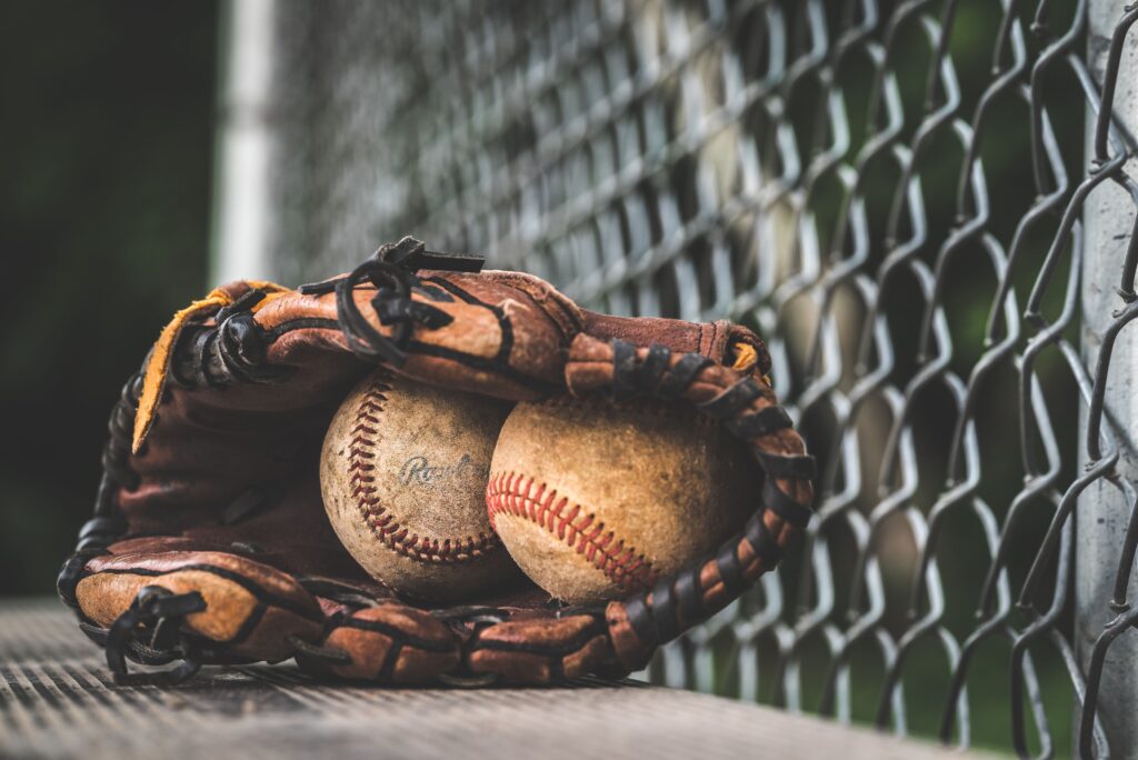 Two worn baseballs in a worn mitt, leaning up against a chainlink fence.
