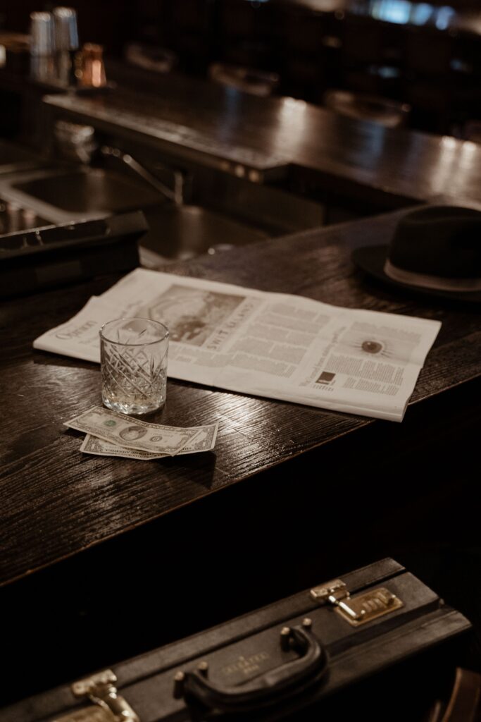 A vintage-style bar interior featuring a black hat, a briefcase, a newspaper, and money on the counter.
