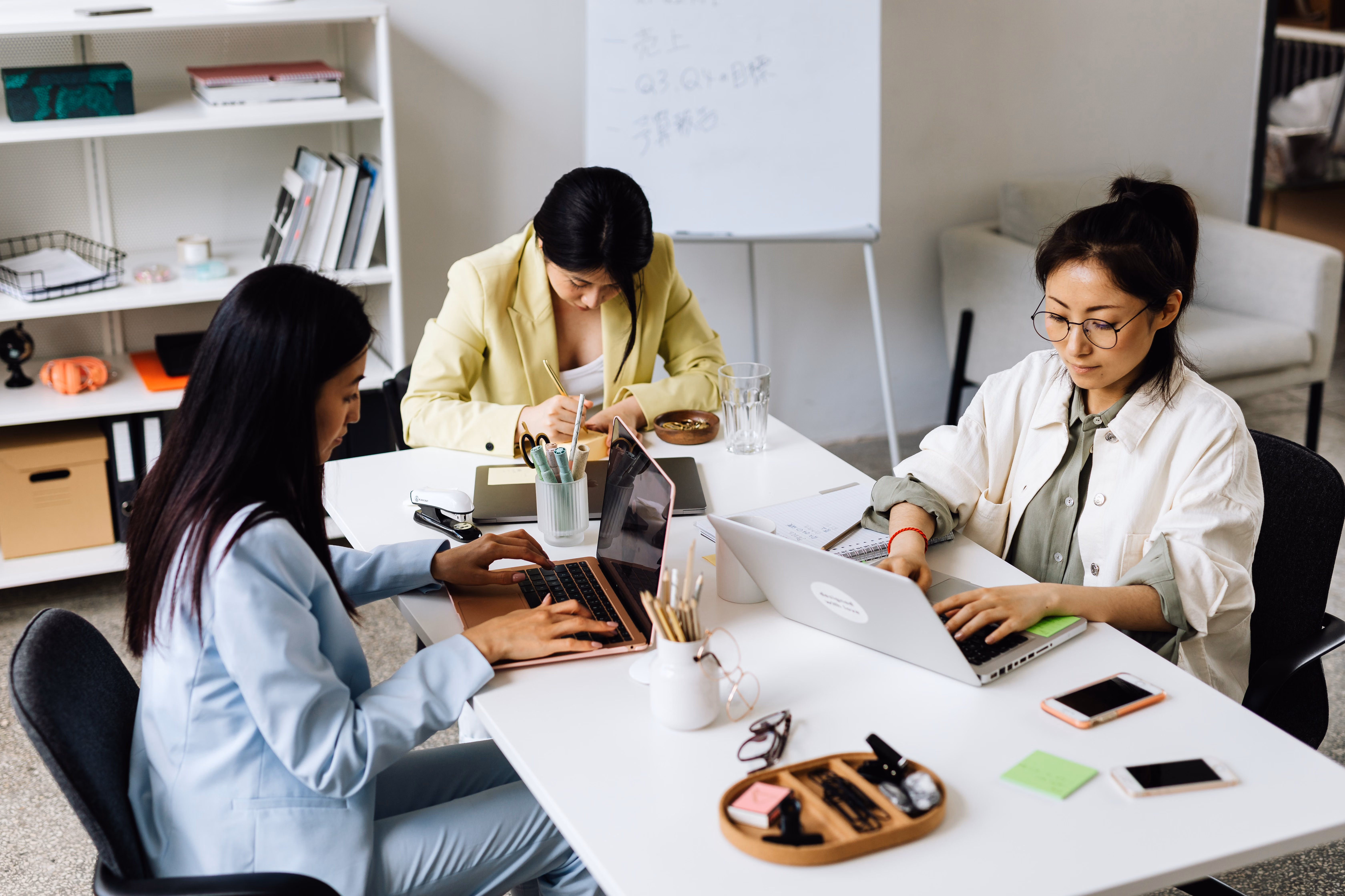 Three Asian women working on laptops together in a modern office.