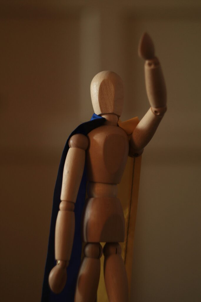 A wooden mannequin, with a deep blue cape tied around their neck, is posed to fly away. Their arm points towards the sky in a heroic pose.