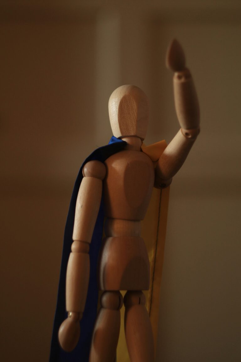 A wooden mannequin, with a deep blue cape tied around their neck, is posed to fly away. Their arm points towards the sky in a heroic pose.