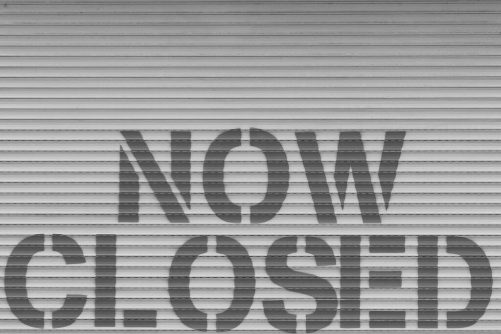 Black & gray image of a 'Now Closed' sign on a roll-down shutter.